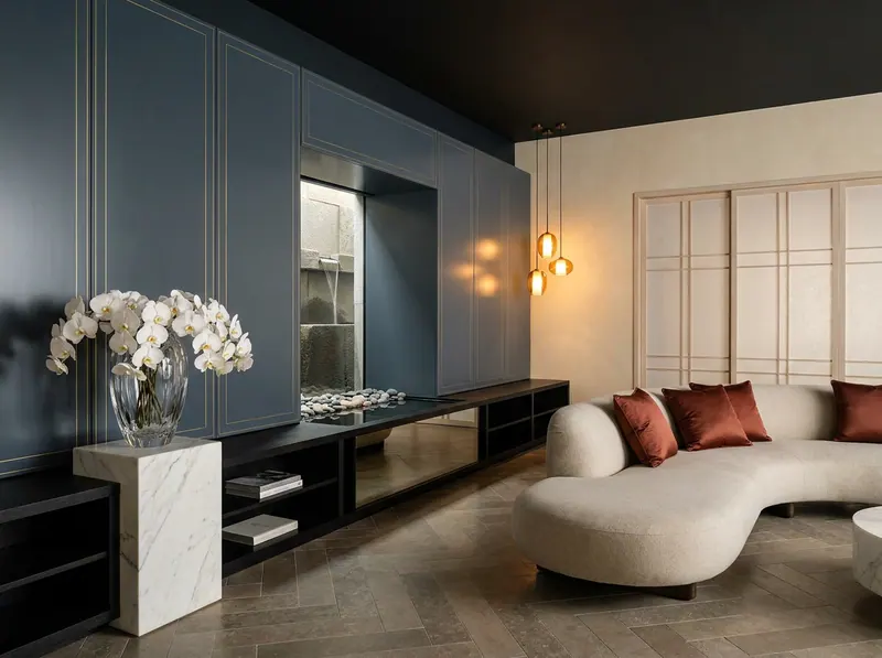 Ultra-luxury East-meets-West fusion interior with slate blue lacquered panels, Japanese stone water feature, crystal glass vessel on marble plinth with white orchids, and terracotta silk cushions — East-meets-West longevity design by Beverly Hills Luxury Interiors